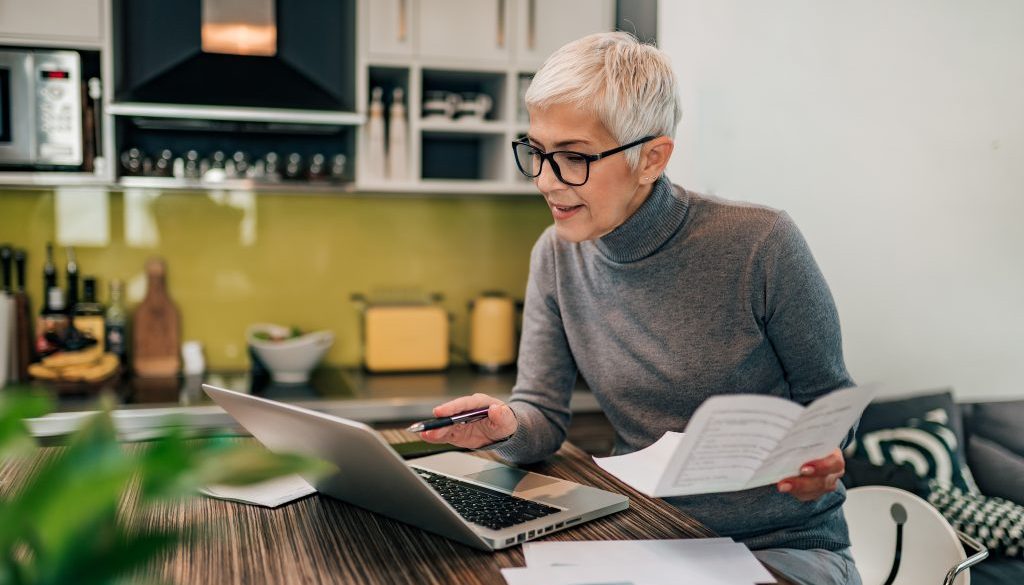 Portrait of a senior woman working with laptop and documents in the kitchen.