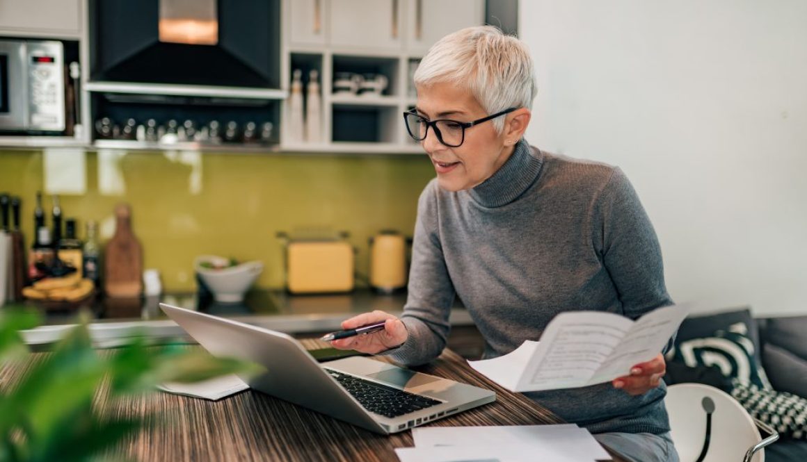 Portrait of a senior woman working with laptop and documents in the kitchen.