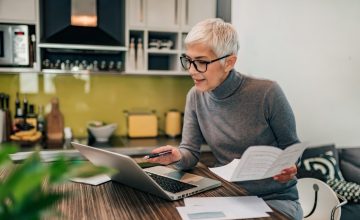 Portrait of a senior woman working with laptop and documents in the kitchen.