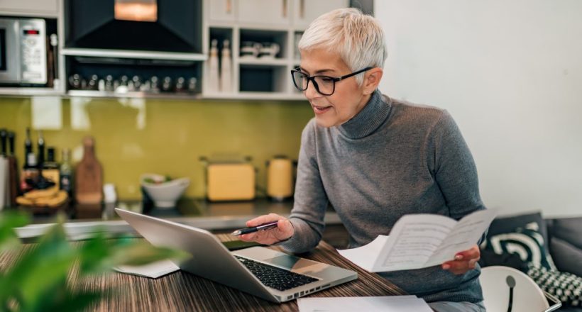 Portrait of a senior woman working with laptop and documents in the kitchen.