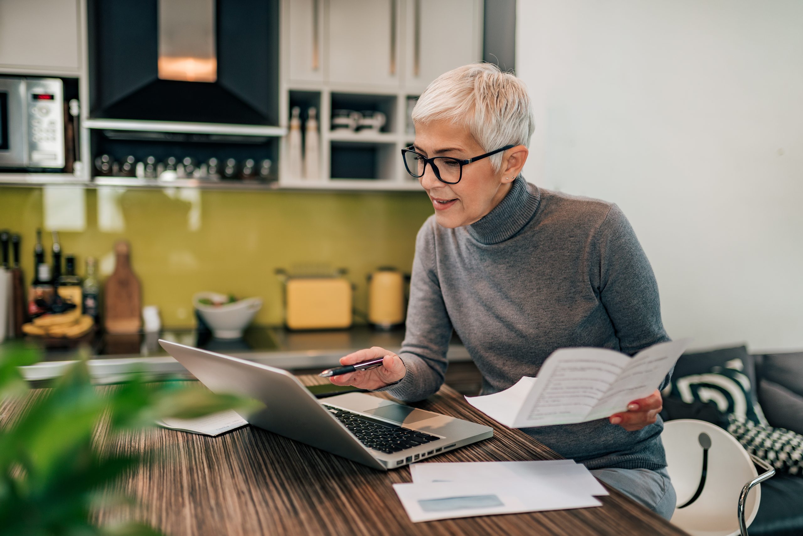 Portrait of a senior woman working with laptop and documents in the kitchen.