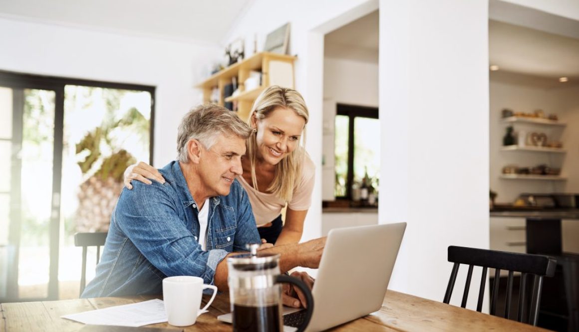 Couple with laptop planning finance, banking and checking retirement budget while becoming debt free at home. Smiling, happy and cheerful mature man showing woman an approved bank loan on technology