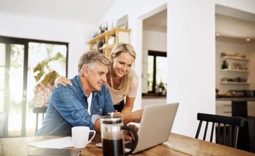 Couple with laptop planning finance, banking and checking retirement budget while becoming debt free at home. Smiling, happy and cheerful mature man showing woman an approved bank loan on technology