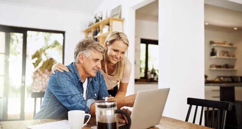Couple with laptop planning finance, banking and checking retirement budget while becoming debt free at home. Smiling, happy and cheerful mature man showing woman an approved bank loan on technology